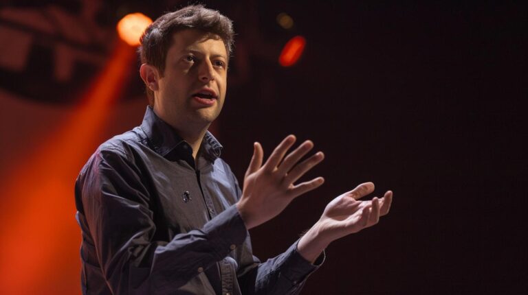 Man speaking at a conference with gesturing hands on stage with red background lights