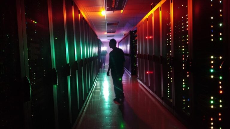 Silhouette of a person walking through a dark data center aisle with colorful blinking lights on server racks