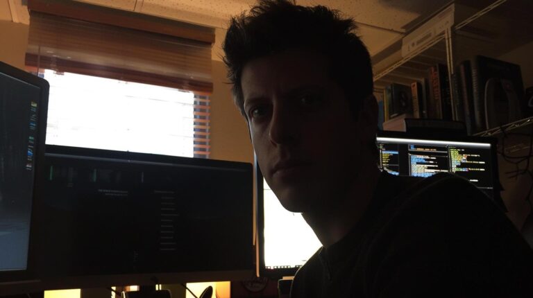 Man sitting in front of computers with code on screens in a dimly lit room.
