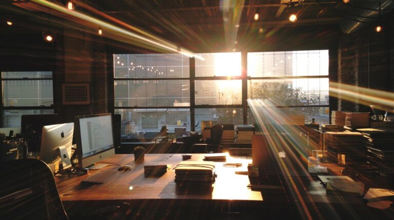 Modern office interior with sunlight streaming through large windows onto desks and computers.
